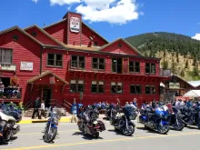 A large, two-story red building housing Texas Reds Steakhouse and The Lodge at Red River, featuring a row of motorcycles parked along the street front. People are walking on the sidewalk and dining on the patio under a bright blue sky with forested mountains in the background.