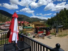 Summer view of the Platinum Chairlift at Red River Ski & Summer Area from a patio deck, featuring a closed red and white umbrella, the lift terminal, and resort lodging against a backdrop of green mountains and blue sky.