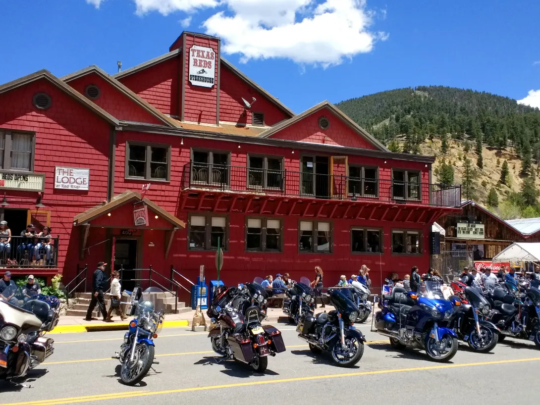 A large, two-story red building housing Texas Reds Steakhouse and The Lodge at Red River, featuring a row of motorcycles parked along the street front. People are walking on the sidewalk and dining on the patio under a bright blue sky with forested mountains in the background.