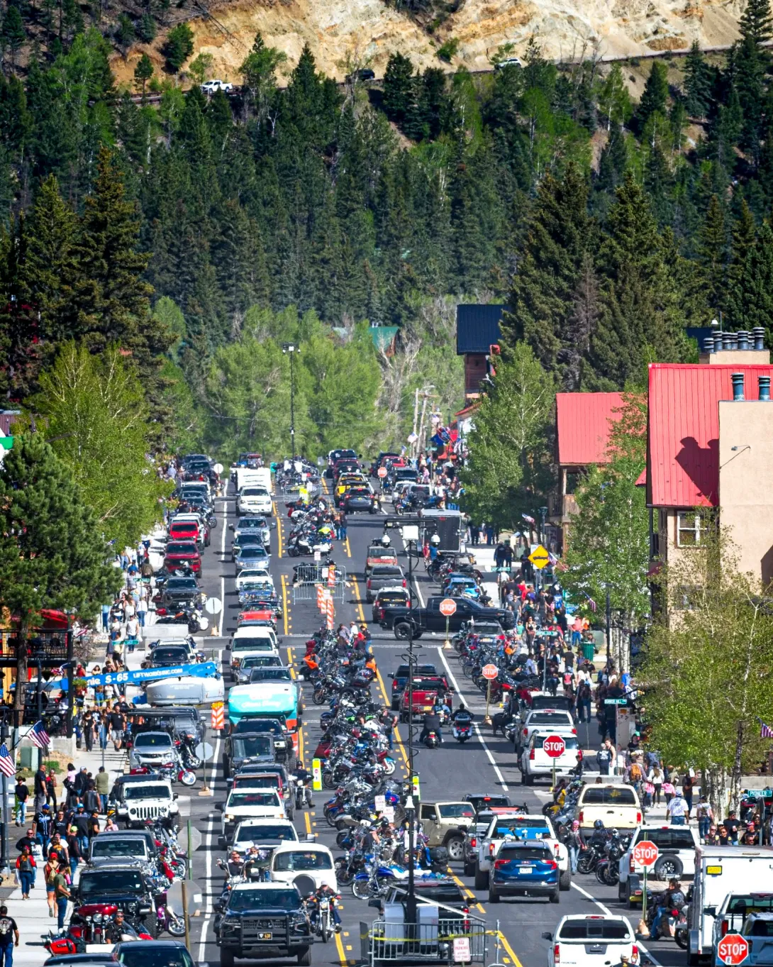A bustling main street in a mountain town heavily congested with traffic, featuring a large number of motorcycles parked down the center median and along the sides of the road. Cars and trucks are moving slowly through the lanes, while dense crowds of pedestrians walk along the sidewalks in front of local businesses. Steep, pine-forested mountains rise in the background.